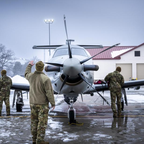 Vier Soldaten in Tarnuniformen inspizieren ein Propellerflugzeug auf einem verschneiten Flugplatz.