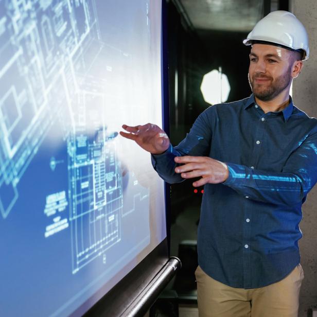 Fachmann im Baustellenhelm zeigt auf digitale Architekturpläne in einem modernen Büro.