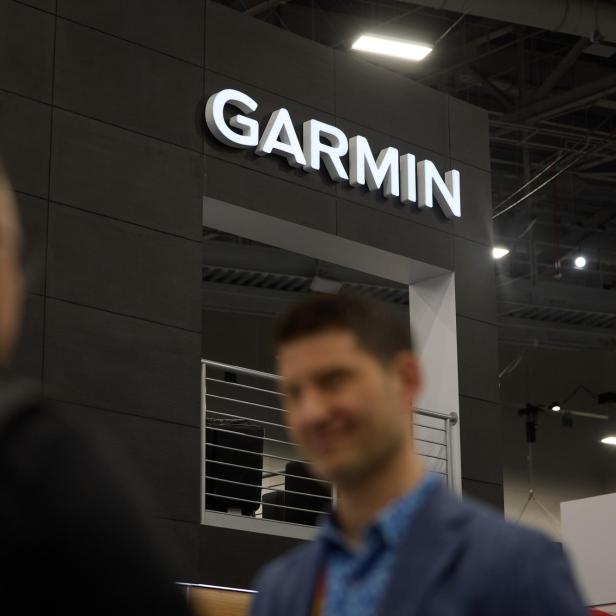 Drei unscharfe Personen stehen vor einem Messestand mit großem leuchtendem Garmin-Schriftzug und moderner Architektur.