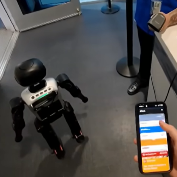 Ein humanoider Roboter steht an einem Flughafenschalter neben einer Person mit Smartphone.
