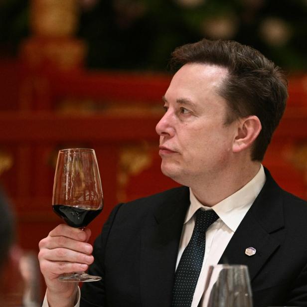 Ein Mann im Anzug hält ein Glas Rotwein und blickt nach links, im Hintergrund rote Dekoration.