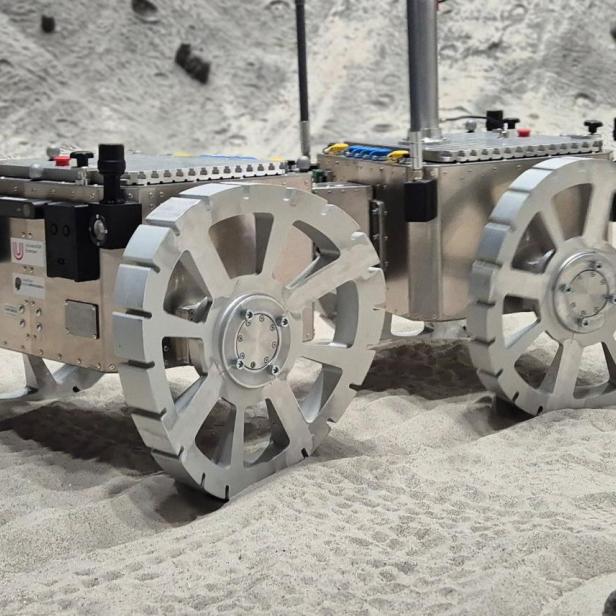 Mars-Rover steht auf sandigem Untergrund des Testgeländes. Seine silbernen Räder sind gebogen.