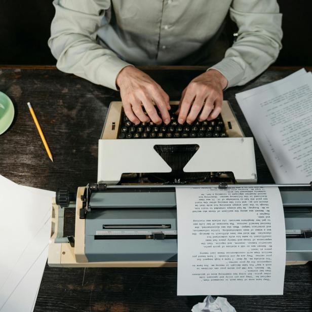 Person tippt an einer Schreibmaschine, umgeben von Papier, Bleistift und Kaffeetasse.