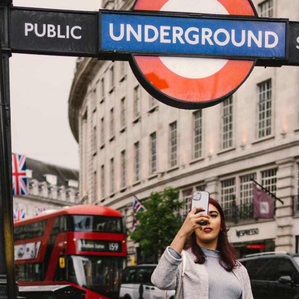 Frau macht ein Selfie vor einem London Underground Schild, im Hintergrund rote Doppeldeckerbusse.