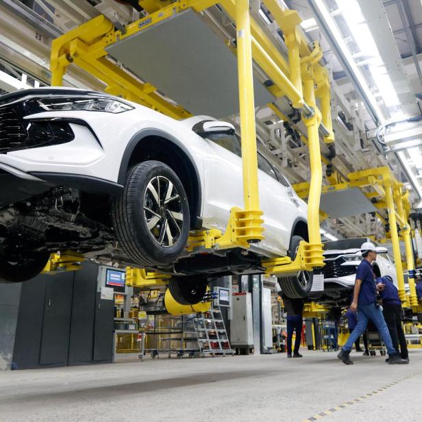 Mehrere weiße Autos hängen an gelben Hebevorrichtungen in einer modernen Autofabrik, während Arbeiter daneben stehen.