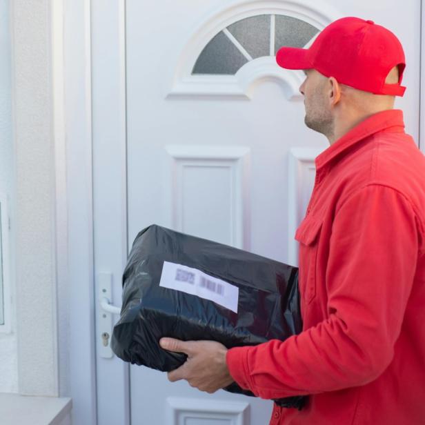 Paketbote in roter Kleidung liefert ein schwarzes Paket an eine Haustür.