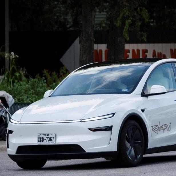 Ein weißes Elektroauto mit schwarzen Felgen und getönten Scheiben fährt auf einer Straße, im Hintergrund sind Fußgänger zu sehen.