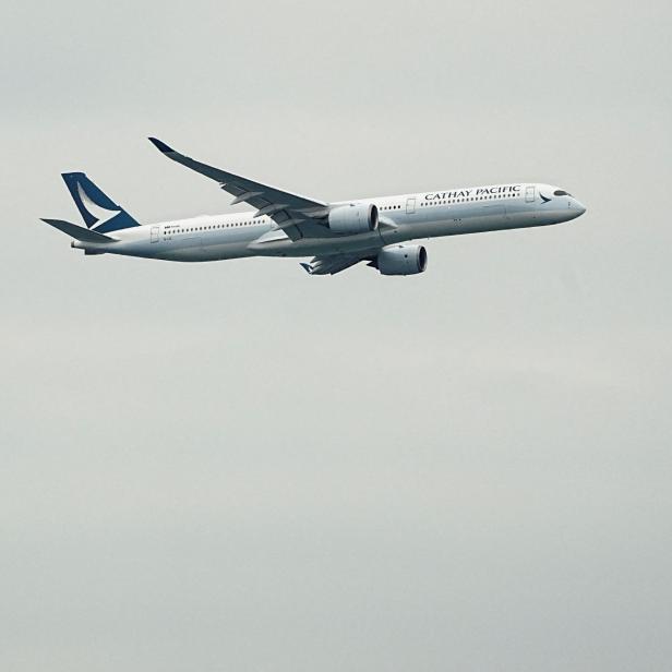 Ein Cathay Pacific Flugzeug fliegt vor einem hohen Gebäude in leicht bewölktem Himmel.