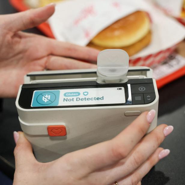 Eine Person hält ein Gerät, das anzeigt, dass in einem Lebensmittel kein Gluten nachgewiesen wurde, im Hintergrund liegt ein Burger.