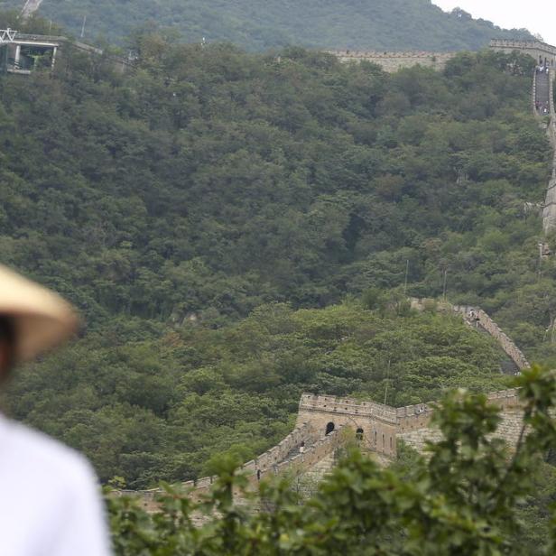 Die Chinesische Mauer schlängelt sich über einen bewaldeten Hügel, im Vordergrund eine Person mit einem traditionellen Hut.