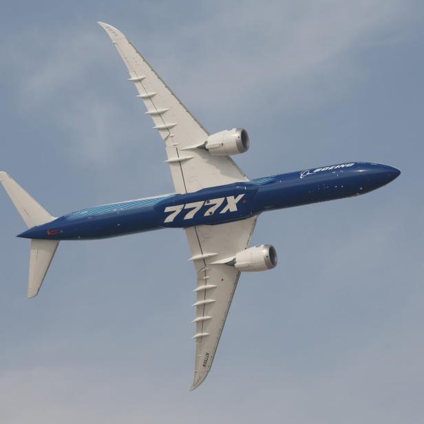 Ein großes blau-weißes Passagierflugzeug mit der Aufschrift „777X“ fliegt schräg am Himmel.