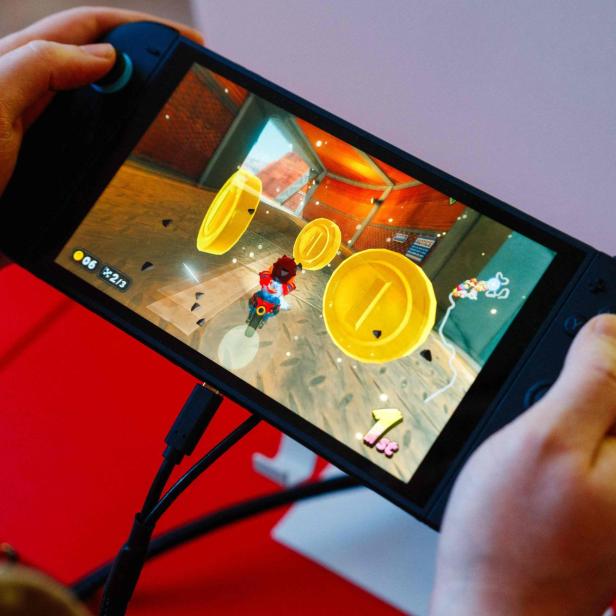Hände halten eine Nintendo Switch, auf der ein Rennspiel mit Münzen und einer „1st“-Platzierung angezeigt wird.