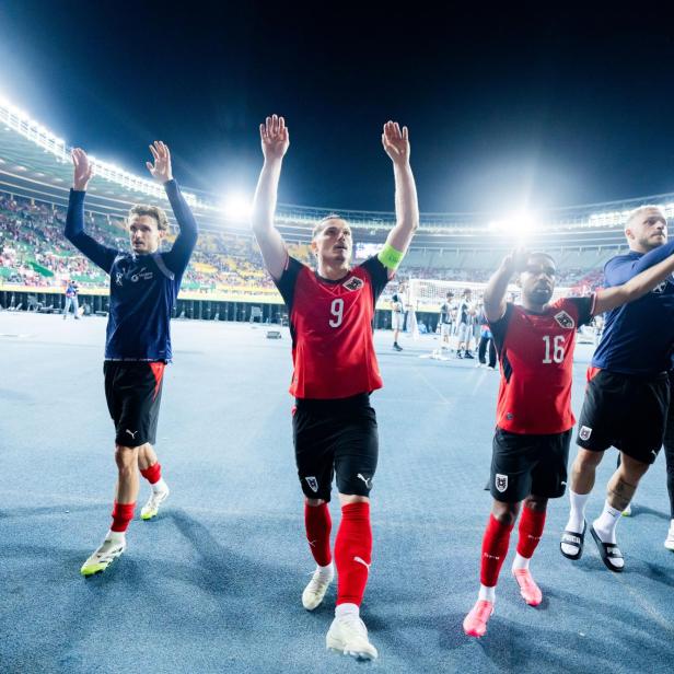 Mehrere Fußballspieler in roten Trikots jubeln mit erhobenen Armen vor vollen Tribünen in einem beleuchteten Stadion.