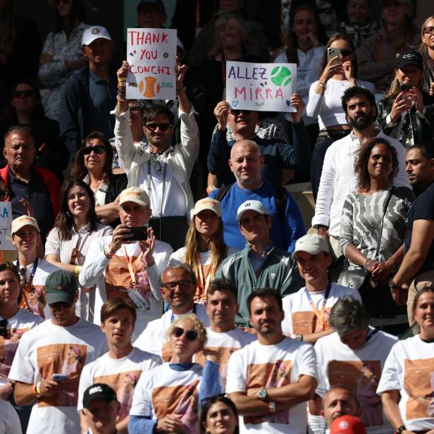 Eine Menschenmenge jubelt, einige halten bunte Schilder mit Tennisbezug hoch, viele tragen weiße T-Shirts mit einem Tennis-Motiv.