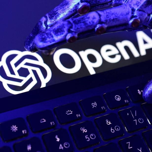 Eine metallische Roboterhand schwebt über einer Tastatur, im Hintergrund ist das OpenAI-Logo zu sehen.