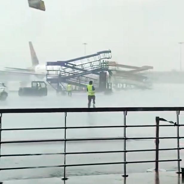 Die Gangway rollt über den Flughafen, das Wetter ist regnerisch und windig. Im Hintergrund steht ein Flugzeug