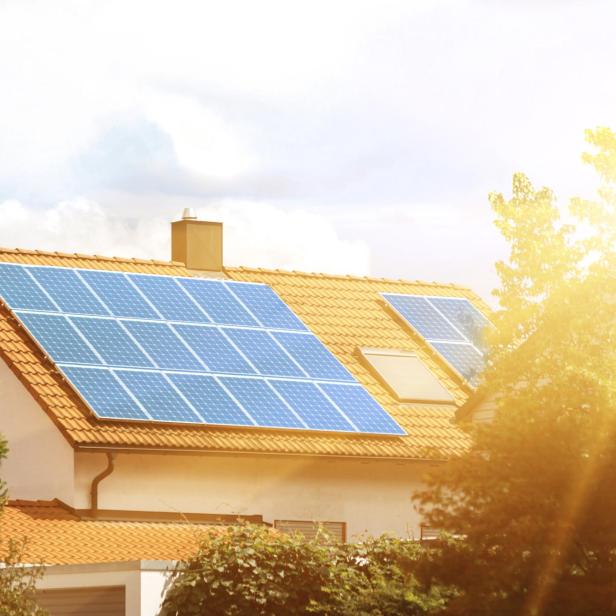 Auf einem Hausdach mit roten Ziegeln sind mehrere Solarpanels installiert, Sonnenstrahlen scheinen darauf.