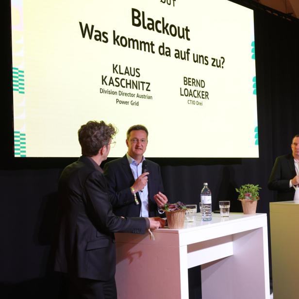 Drei Männer im Anzug diskutieren auf einer Bühne mit Mikrofonen vor einer großen Präsentationsfolie zum Thema Blackout.