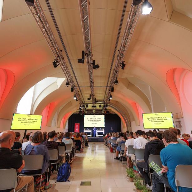 Viele Menschen sitzen in einem modernen Saal mit gewölbter Decke und hören einem Vortrag auf einer Bühne mit großen Bildschirmen zu.