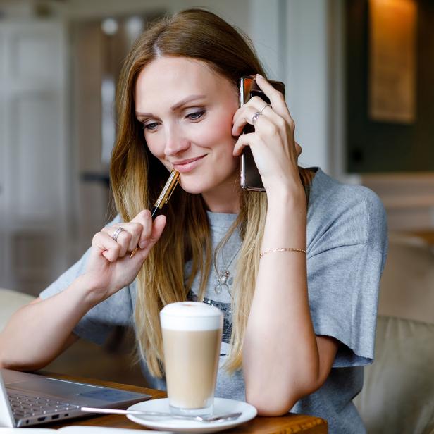 Eine Frau sitzt vor dem Notebook und telefoniert am Smartphone.