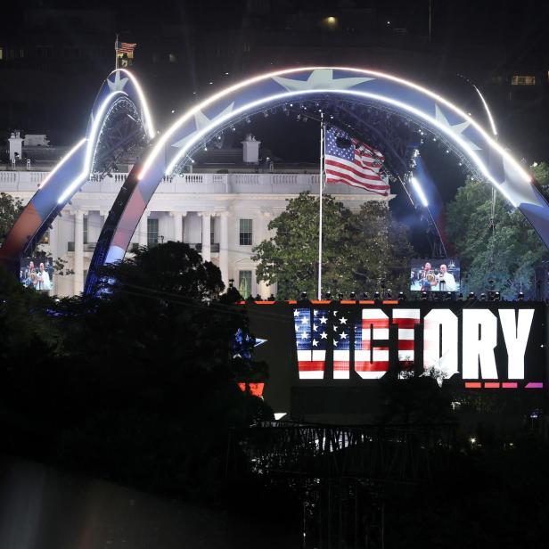 Große Bühne mit leuchtendem Schriftzug „VICTORY“, US-Flagge und dem Weißen Haus im Hintergrund bei Nacht.