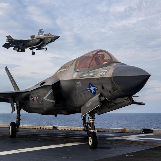 Zwei US-Kampfjets vom Typ F-35 befinden sich auf einem Flugzeugträger, einer steht auf dem Deck, der andere landet gerade.