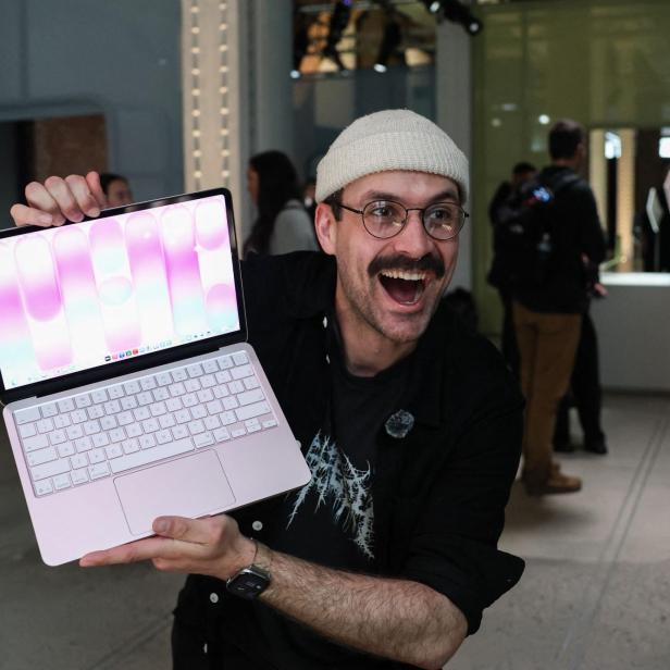 Ein Mann mit Mütze und Brille hält ein rosa Laptop hoch und grinst begeistert in einem Ausstellungsraum.