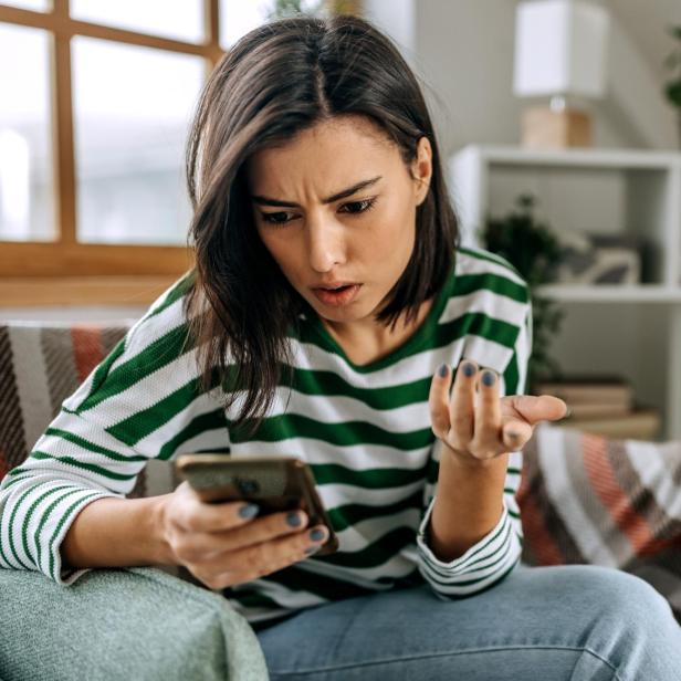 Eine Frau schaut irritiert auf ihr Smartphone und gestikuliert fragend auf dem Sofa.