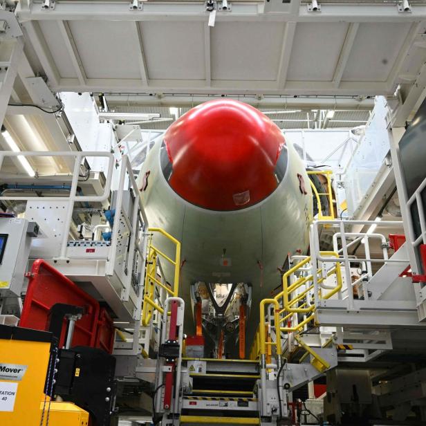 Flugzeugrumpf mit roter Nasenverkleidung in einer Industrieanlage, umgeben von Arbeitsbühnen und Maschinen.
