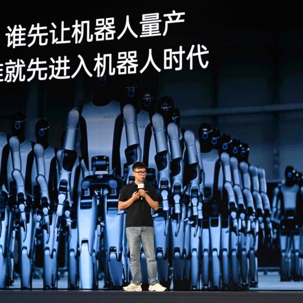 Vortragender vor riesiger Robotergruppe mit chinesischer Schrift auf der Leinwand.