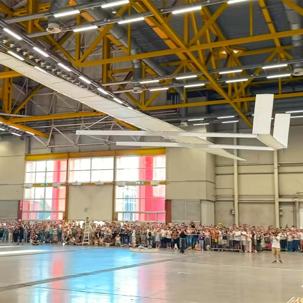 Ein riesiger Papierflieger wird in einer Halle mit Publikum gestartet.