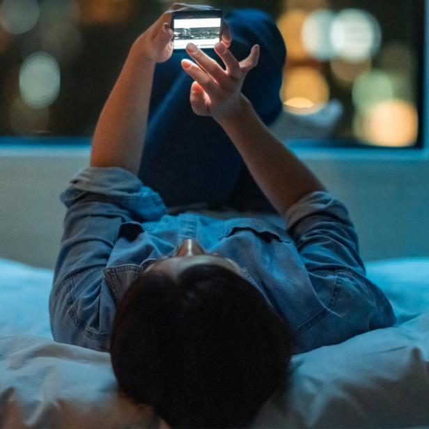 Person liegt auf dem Bett und hält ein Smartphone hoch.