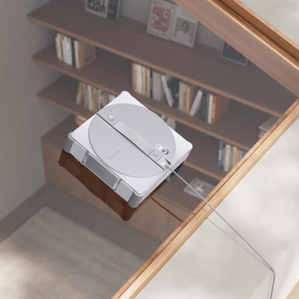 Ein ECOVACS Winbot W3 Omni Fensterputzroboter reinigt eine Glasfläche über einem Bücherregal.