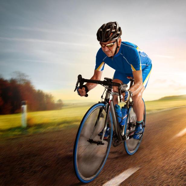 Ein Rennradfahrer in blauer Sportkleidung und mit Helm fährt bei Sonnenuntergang auf einer Landstraße.
