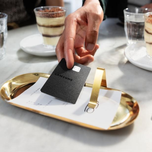 Schwarze Samsung Kreditkarte liegt auf einem goldenen Tablett mit der Rechnung, im Hintergrund stehen Gläser mit Dessert und Wasser