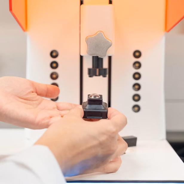 Hände halten eine kleine Linse vor einem 3D-Drucker.