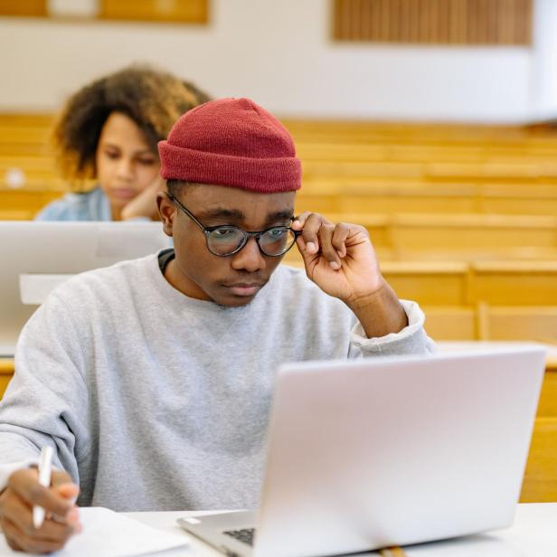 Studierender mit roter Mütze arbeitet in einem Hörsaal am Laptop, während hinter ihm eine Kommilitonin ebenfalls mit Computer sitzt.