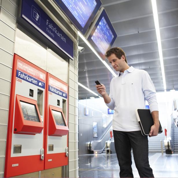 Ein Mann steht an einem Fahrkartenautomaten und benutzt sein Smartphone.