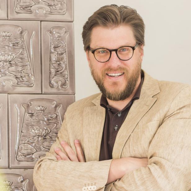 Ein Mann mit Brille und beigem Sakko steht vor einem Kachelofen.