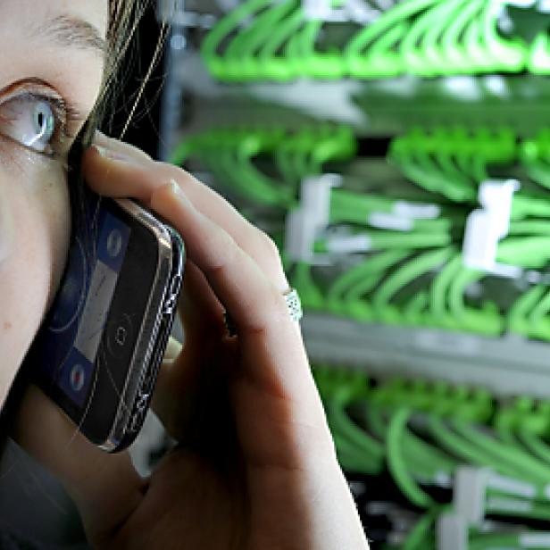 Eine junge Frau telefoniert vor einem Serverschrank mit grünen Kabeln.